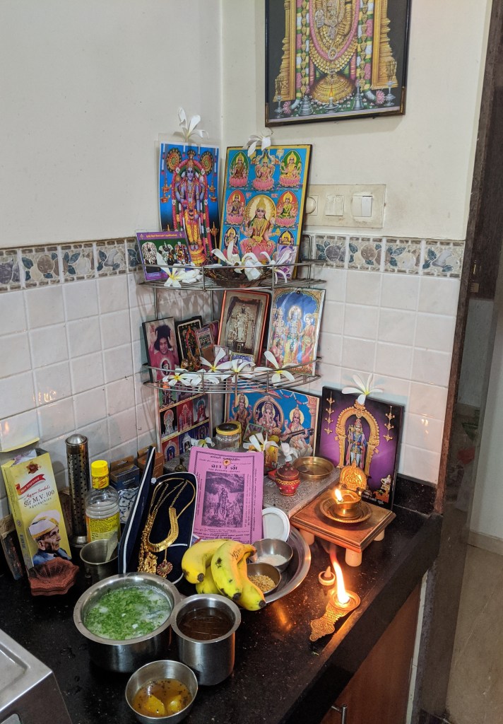 Prayer altar with photos of deities a lamp burning. Offerings of jewels, fruits, rice, dal, currency and coins. Along with neer mor, panakand a mango, neem flower and jaggery mix.